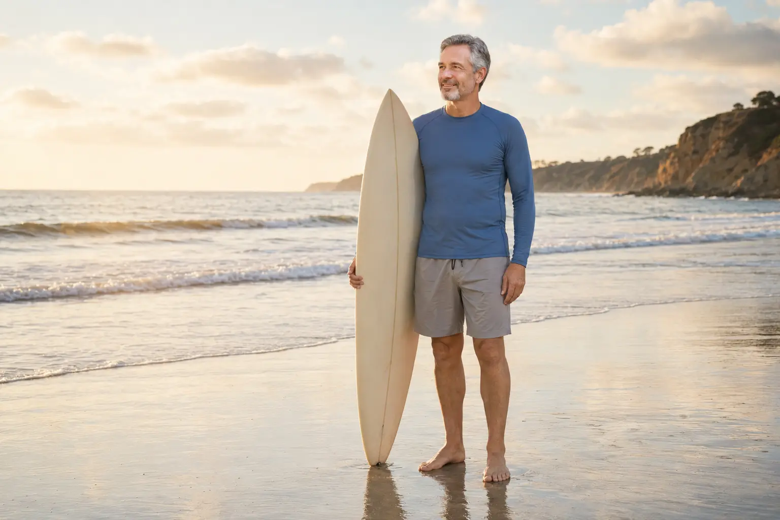 Man in his 50s standing comfortably on a beach with a surfboard