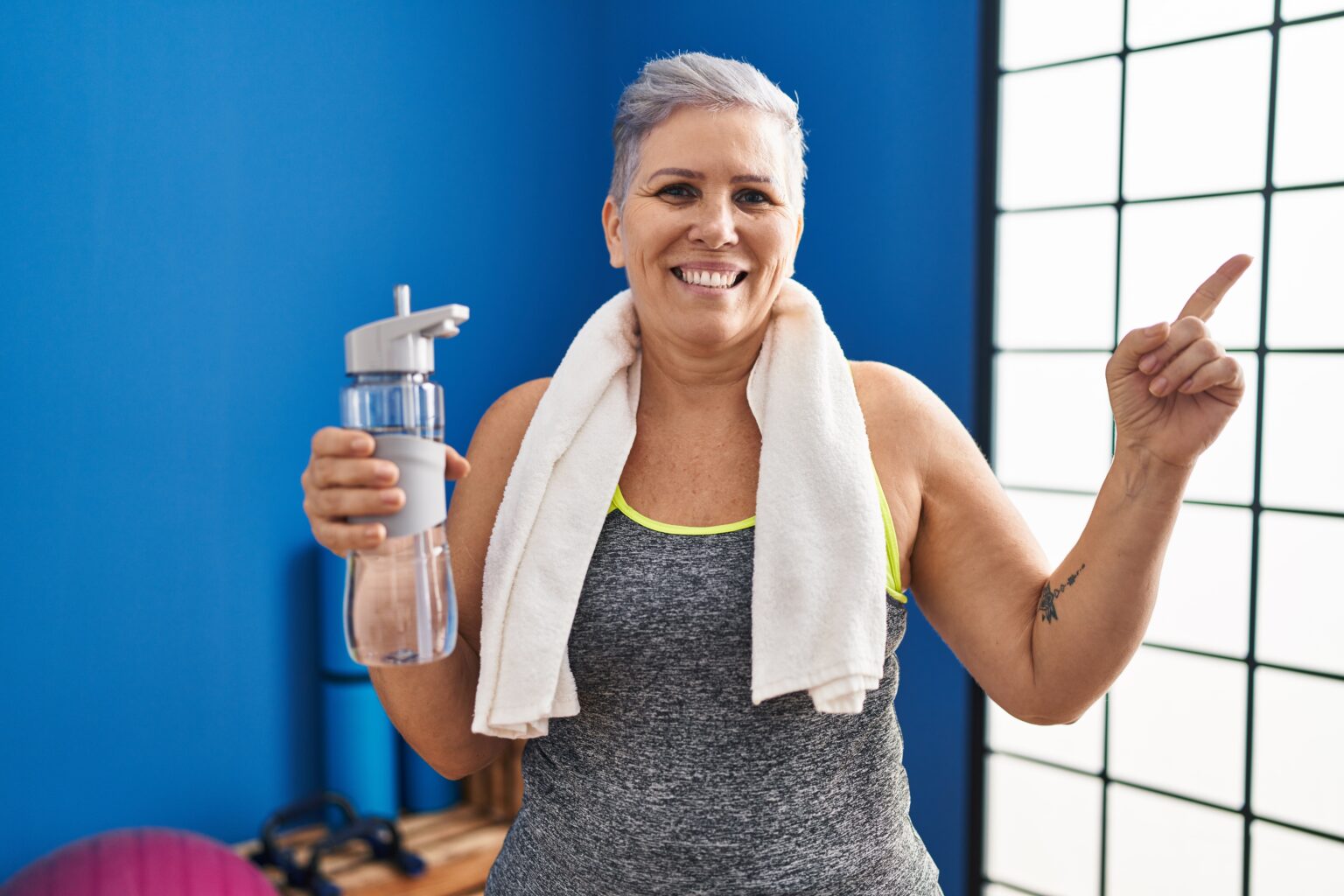 Middle age woman wearing sportswear and towel holding water smiling happy pointing with hand and finger to the side